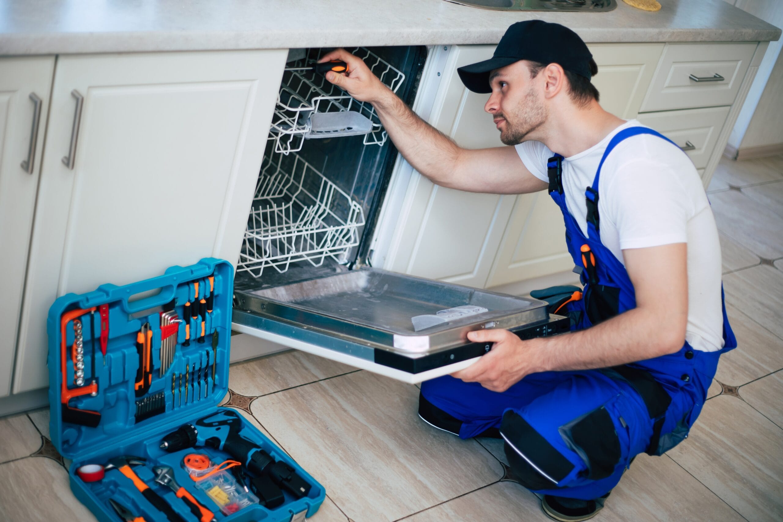 Young modern serviceman in worker suit during the repairing of the dishwasher on the domestic kitchen.